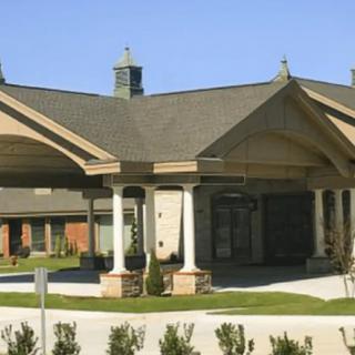 A building with a prominent entrance featuring large columns and a sloped roof, surrounded by landscaped greenery.