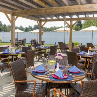 An inviting outdoor dining area with neatly arranged tables and chairs under a wooden pergola, featuring colorful napkins and decorative flowers.
