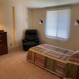 A cozy bedroom with a single bed, a recliner chair, a dresser, and natural light coming through the window.
