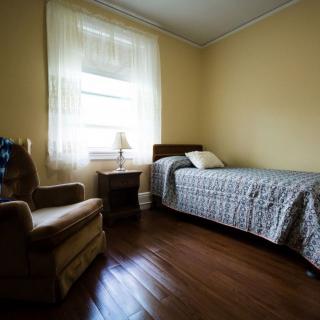 A simple bedroom with a single bed, an armchair, and a window letting in natural light.