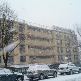A snowy day in the city, with cars parked on the street and a multi-story building partially obscured by falling snow.