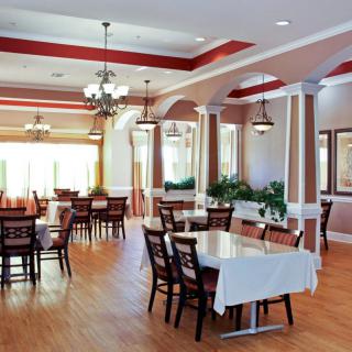An elegant dining area featuring tables with white tablecloths, wooden chairs, chandeliers, and decorative columns.