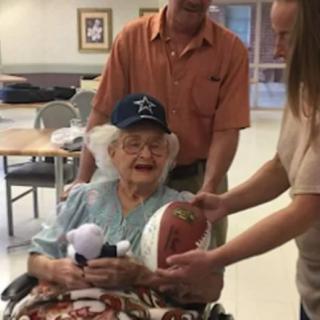 An elderly woman in a wheelchair smiles as she holds a football, surrounded by two people indoors.