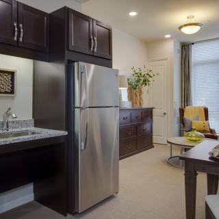 A modern kitchen and living area in an apartment, featuring a stainless steel refrigerator, dark wood cabinets, and comfortable seating.