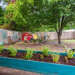 A vibrant outdoor garden area featuring raised beds with plants, a colorful play structure, and a large tree providing shade.