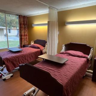 A hospital room with two beds separated by a curtain, featuring large windows and tray tables.