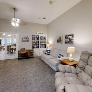 A cozy living room with sofas, armchair, and a ceiling fan, featuring warm lighting and wall decorations.