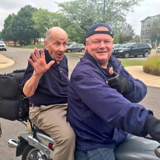 Two men enjoying a motorcycle ride together, smiling and waving.