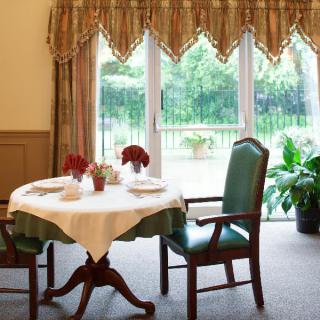 A cozy dining area with a neatly set table by a large window, adorned with elegant curtains and surrounded by greenery.