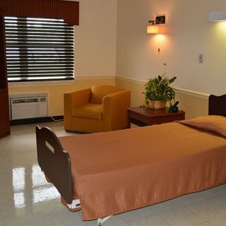 A hospital room with a single bed, a chair, and a small table near a window.