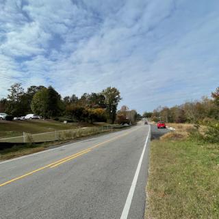 A rural road with cars and trees under a partly cloudy sky.