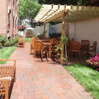 A cozy outdoor patio with wooden furniture and a pergola, set alongside a brick pathway and surrounded by greenery.