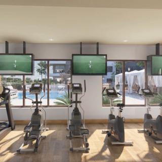 A modern gym with stationary bikes and a treadmill, featuring multiple televisions and a scenic pool view through large windows.