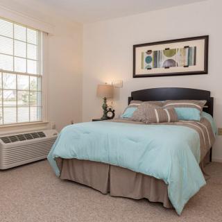 A cozy bedroom featuring a neatly made bed with blue bedding, a bedside lamp, and a large window allowing natural light.