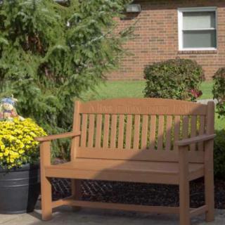 A wooden bench in a garden setting next to yellow flowers and shrubs, with a brick building in the background.