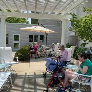 A sunny outdoor patio with people sitting, some using wheelchairs and walkers, enjoying a pleasant day.