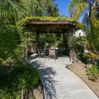 A tranquil garden scene featuring a pergola with outdoor seating, surrounded by lush greenery and tropical plants.