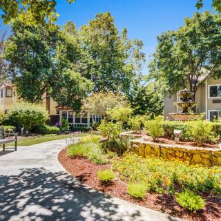 A serene garden pathway surrounded by lush greenery and residential buildings on a sunny day.