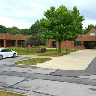 A view of a brick building surrounded by greenery and a parked white car.