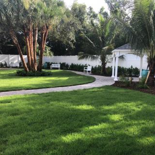 A beautifully landscaped garden with a winding pathway, surrounded by a lush green lawn, palm trees, and a charming gazebo.
