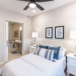 A modern bedroom with a ceiling fan, featuring a neatly made bed with blue and white pillows, two nightstands with lamps, and an adjacent bathroom.