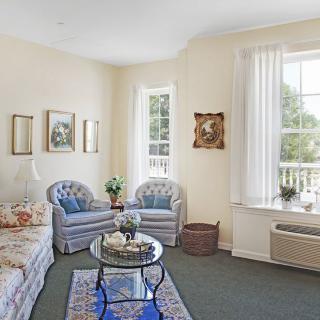 A cozy living room with elegant furniture, featuring a sofa, chairs, and a glass coffee table, accented by artwork on the walls and large windows.