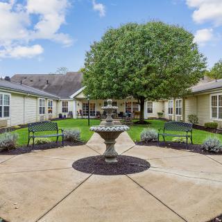 A peaceful courtyard featuring a fountain, surrounded by benches and greenery, with buildings in the background under a blue sky.