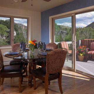 A cozy dining room with a glass table and upholstered chairs, overlooking a scenic mountain view through large windows and sliding doors leading to a deck.