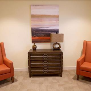 A cozy sitting area featuring two orange chairs, a wooden chest with decorative items, and a colorful abstract painting above.