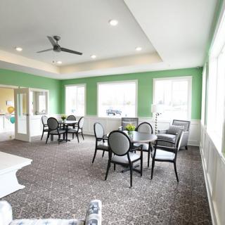 A brightly lit dining area with green walls, featuring several tables and chairs, carpeted flooring, and large windows.