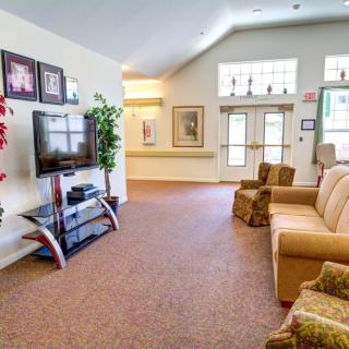 A cozy living room with a sofa, armchairs, and a television set against a wall decorated with framed photos, complemented by large windows and indoor plants.