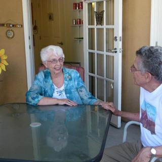 An elderly woman and a man having a conversation at a glass table on a patio.