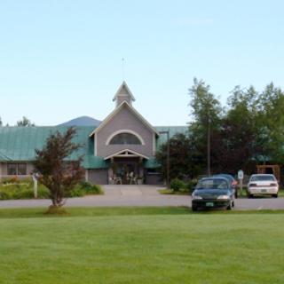 A scenic view of a building with a green roof, surrounded by trees and parked cars on a grassy lawn.
