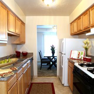 A modern apartment kitchen with wooden cabinets and white appliances, leading to a dining area.