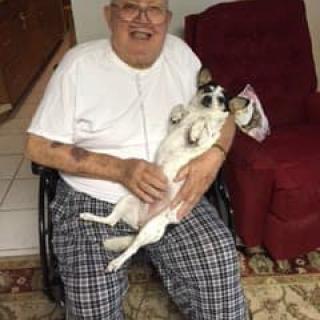 A man sitting in a wheelchair, smiling and holding a small dog indoors.