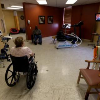 Elderly individuals in wheelchairs are seated in a healthcare facility room equipped with rehabilitation equipment.