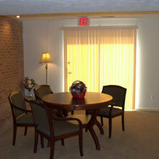 A cozy dining area with a round table and chairs, softly lit by a floor lamp beside window blinds.
