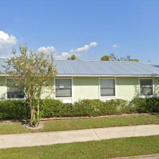 A single-story house with solar panels on the roof, surrounded by a well-maintained lawn and shrubbery under a clear blue sky.