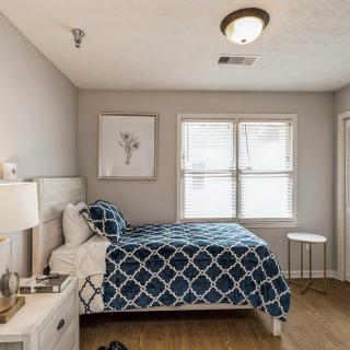 A cozy bedroom with a neatly made bed featuring a blue geometric-patterned duvet, flanked by a nightstand with a lamp, and artwork hanging on the wall. The room has ample natural light coming through the window.