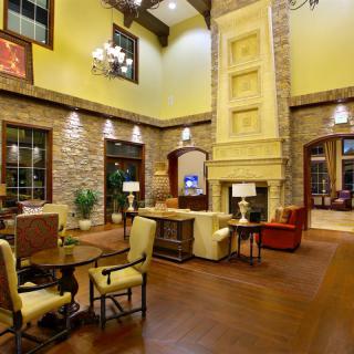A warm and inviting living room featuring a tall stone fireplace, wooden floors, and elegant furnishings under a high ceiling with a chandelier.