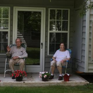 A couple sitting on chairs outside their house, surrounded by potted flowers.