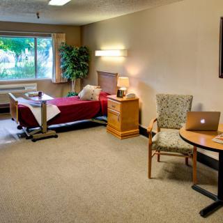 A well-furnished hospital room featuring a bed, table, chair, and large window.