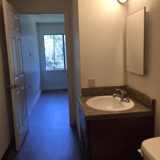 A bathroom with a sink and mirror, leading to a room with a window.