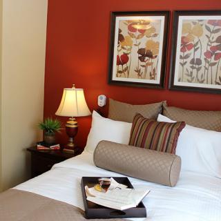 A cozy bedroom with warm red walls, decorative pillows, wall art, and a bedside lamp.
