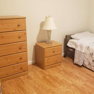 A simple bedroom with a wooden chest of drawers, nightstand with a lamp, and a bed.