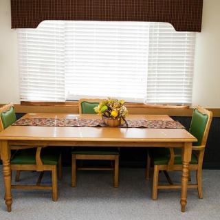 A dining area with wooden table and green-cushioned chairs, featuring a centerpiece on the table in front of a window with blinds.