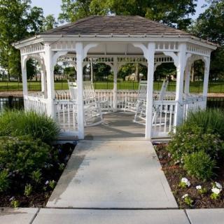 A charming white gazebo surrounded by lush greenery, with a pathway leading to its entrance in a serene park setting.