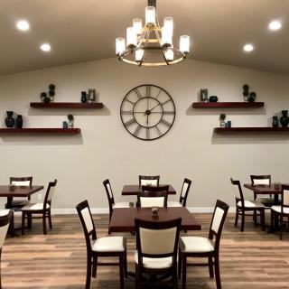 A cozy dining area with neatly arranged tables and chairs, featuring wall shelves, a large clock, and a chandelier.