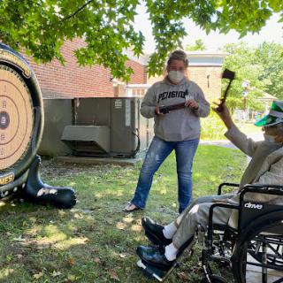 A person in a wheelchair participates in an outdoor archery activity, preparing to throw a hatchet at a target while another person observes.