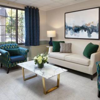 A cozy living room with a cream sofa, two patterned armchairs, and a marble top coffee table adorned with flowers. The space is complemented by abstract wall art, a lamp, and large windows framed by dark curtains.